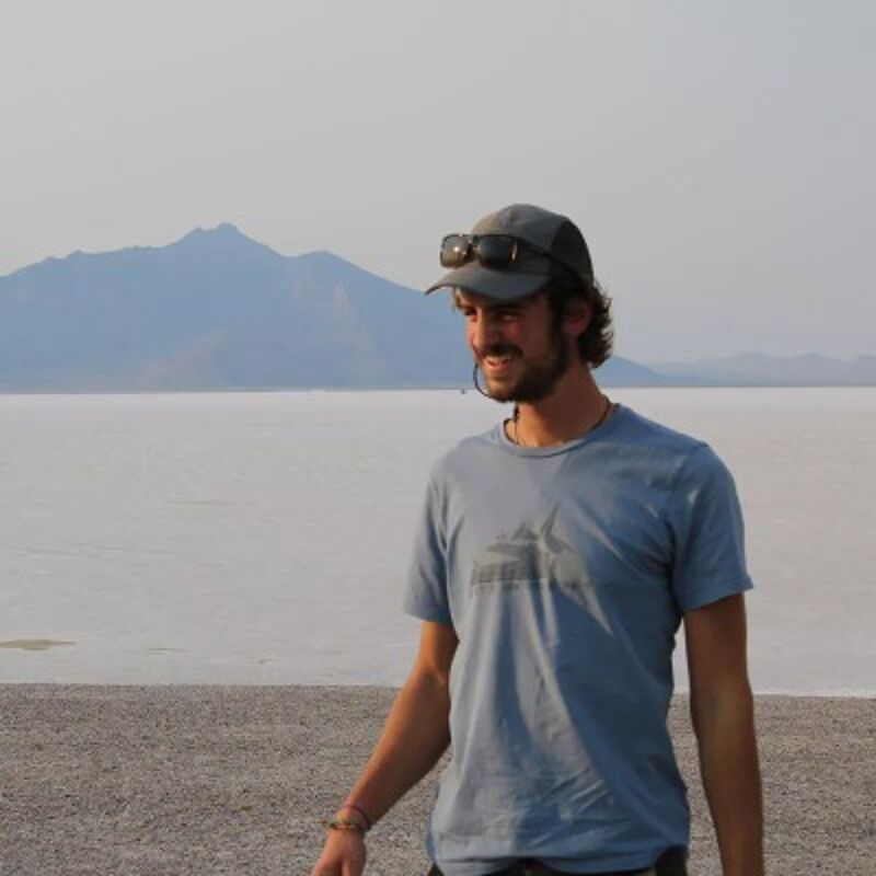 The image shows a man standing in front of a large, flat, white expanse, likely a salt flat or a similar environment. He is wearing a blue t-shirt, a cap with sunglasses perched on top, and appears to be smiling. In the background, there's a mountain range under a hazy sky.